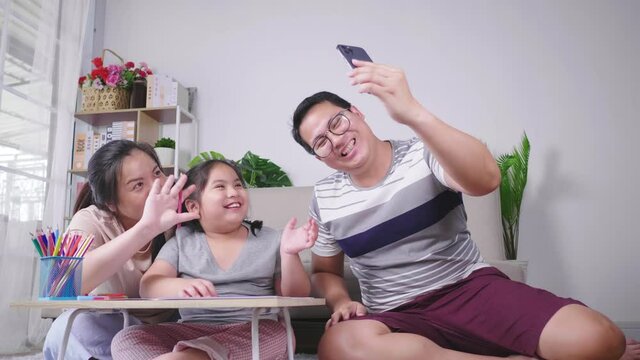 Asian family taking video call on dad's phone during kid painting on paper with mom in living room, sitting on the ground in living room. Having family activity on the weekend. Happy family concept