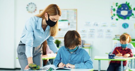 Portrait of Caucasian little junior student writing in copybook studying in classroom listening to...