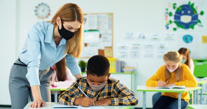 Portrait of young beautiful Caucasian woman teacher helping little African American junior student boy to write test in copybook at class. Female teaching bright schoolboy explaining lesson Post-covid