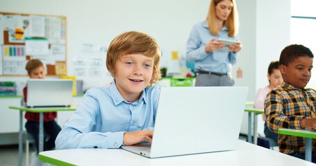 Close up of Caucasian little primary school student sitting at desk in classroom typing on laptop at lesson, female teacher walking behind using tablet. Mixed-race pupils learning at computer class