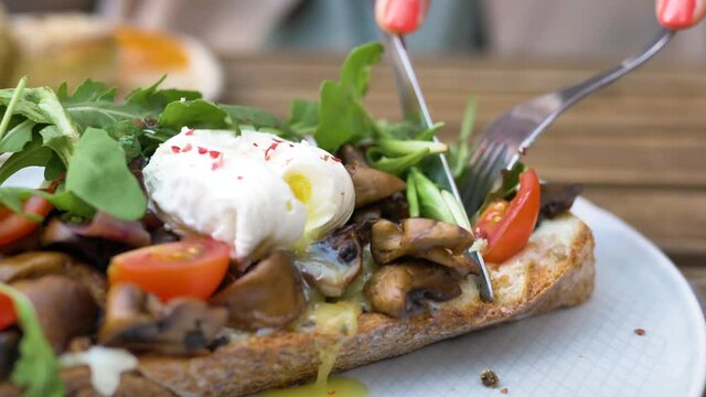 Woman hands cut canape with pouched egg mushrooms tomatoes and ruccola on piece of bread with knife and fork extreme closeup