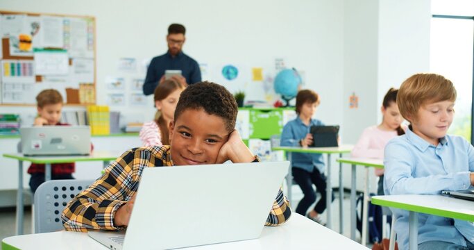 Close up portrait of happy little african american boy pupil using laptop at lesson. Male teacher explaining using tablet on background. Internet technology. Computer science class. Modern education