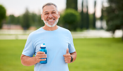 Happy positive mature man with broad smile holding water bottle while doing sport in city park