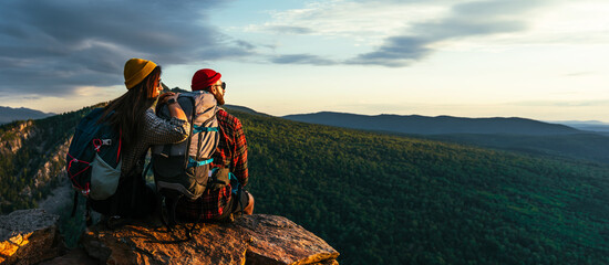 The couple meets the sunset in the mountains. Two travelers are sitting on the edge of a cliff and...