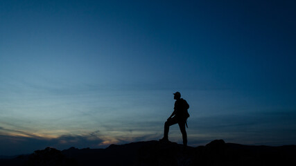 Silhueta de um homem no alto de uma montanha admirando a paisagem logo após o por do sol. fundo azul com espaço para texto.