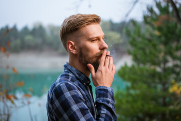The bearded man smokes. Handsome stylish man in plaid shirt with cigarette. Nicotine addiction and bad habits. Caucasian guy stands in the evening by the lake in the forest