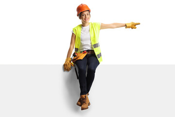 Female construction worker wearing a hardhat and a tool belt sitting on a panel and pointing