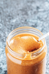 Spoon dipping into a glass jar with crunchy peanut butter. Place for text. Food background.