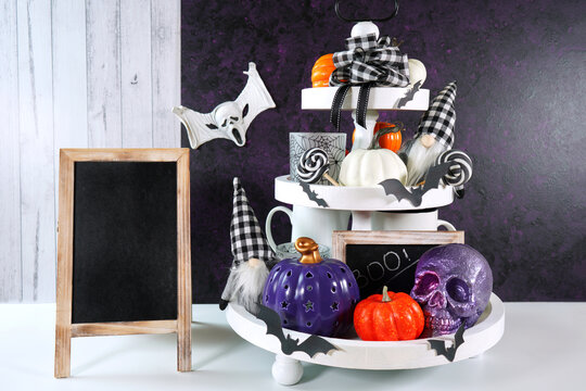 Halloween Farmhouse 3 three tier tray decorated with purple, white and orange pumpkins, skull, lollipops and black plaid gnomes. Farmhouse wooden sign board with copy space.