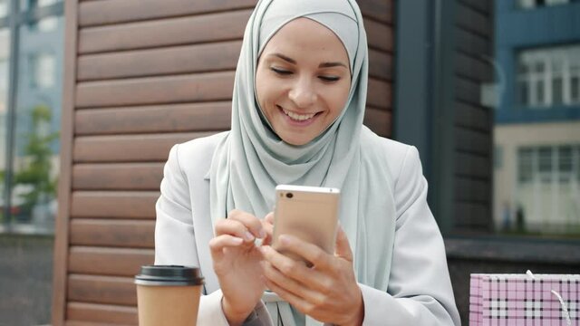 Slow motion of cute Arab woman in hijab using smartphone smiling and drinking coffee in street cafe. Modern lifestyle and outdoor fun concept.