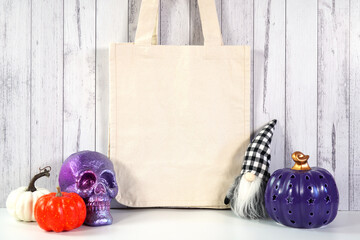 Halloween farmhouse theme tote bag mockup with purple skull, prple orange and white pumpkin, and black plaid gnome against a white wood background Product mock up with negative copy space.