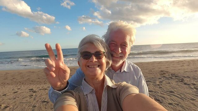 Portrait of couple of mature and old people enjoying summer at the beach looking to the camera taking a selfie together with the sunset at the background. Two active seniors traveling outdoors.
