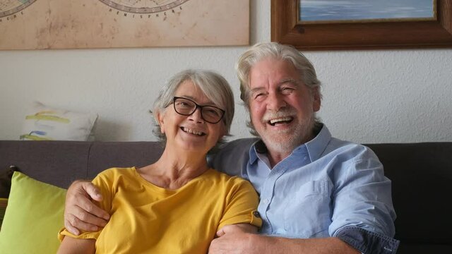 Portrait of couple of seniors laughing and having fun together sitting on the sofa of home indoor. Two old and mature people smiling and enjoying joking.
