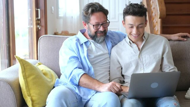 Happy Young Father Sit On Couch Using Laptop Relax With Student Son Holding Smartphone Have Fun Together, Smiling Dad And Little Teenager Boy Enjoy Weekend At Home Rest On Sofa Busy With Gadgets
