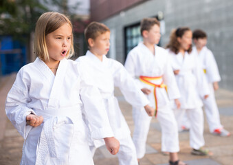 Fototapeta na wymiar Focused preteen girl mastering new karate moves during group class in courtyard of sports school on summer day..