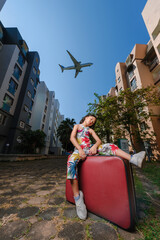 A good looking Asian girl about 8years old wearing a vintage dress posing for a photo with an old red vintage suitcase alone with a large plane flying in the background.