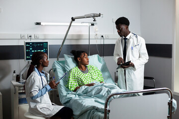 Afro doctors consulting young adult in hospital ward at healthcare clinic. African american people talking to patient wearing cervical collar for neck while sitting in bed. Medical team