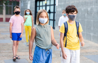Preteen schoolchildren boy and girl in face masks walking home outside school building after finishing lessons on summer day