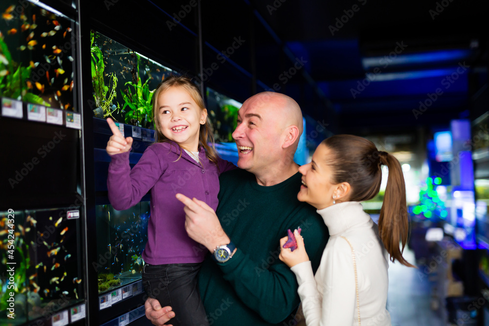 Wall mural Cheerful parents with little girl in arms looking for aquarium fish in pet store