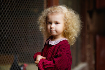 Selective focus. A little blonde girl in a burgundy knitted sweater at the zoo near a cage with birds.