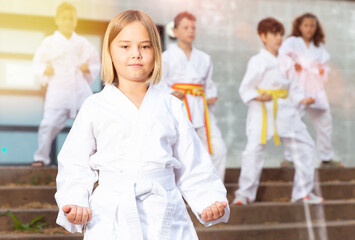 Fototapeta na wymiar Cute schoolgirl in white kimono training karate at schoolyard together with her friends