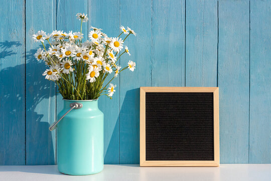 Holiday mockup. Empty blank black letter board and bouquet of natural chamomile in tin can vase on table against blue wooden wall. Front view, Mockup