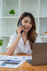 Confident Asian woman talking on her mobile phone and using laptop while sitting at desk and working from home. Home office.