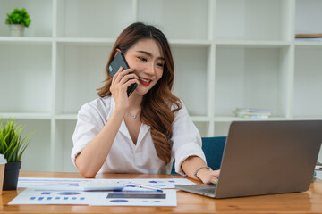 Confident Asian woman talking on her mobile phone and using laptop while sitting at desk and working from home. Home office.