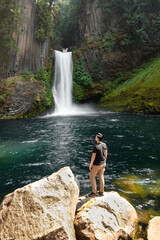 View of a person standing in front of a waterfall in Oregon. Toketee Falls can be seen with cascading water and green moss in a lush forest with a small turquoise pond.