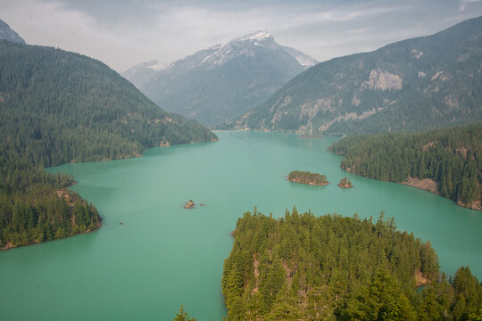 View of diablo lake in North Cascades National Park. A mountain peak with snow can be seen as well as a hazy sky and a forest of green trees.