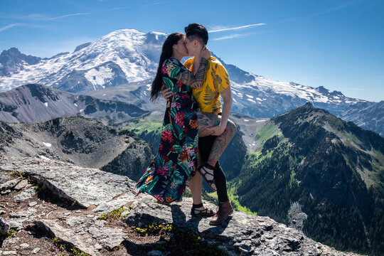 View of lesbian couple kissing in front of Mount Rainier in Mount Rainier National Park. The peak of the mountain is covered in snow and below is fields of green grass.