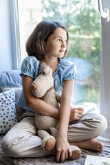 Portrait of a cute pretty teenage girl. The girl sits on the windowsill with her favorite soft toy teddy bear. Soft focus. Positive emotions. Relax and rest. Vertical shot