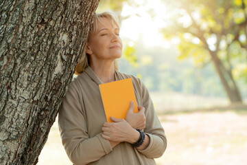 Portrait of happy senior woman relax and hug book under tree in park