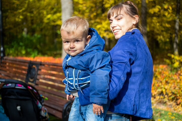 Fototapeta na wymiar Smiling baby boy with dirt on his face looks at the camera and sits in mother arms. woman in blur looks at her son and smiles. close-up, soft focus. in the background autumn golden trees