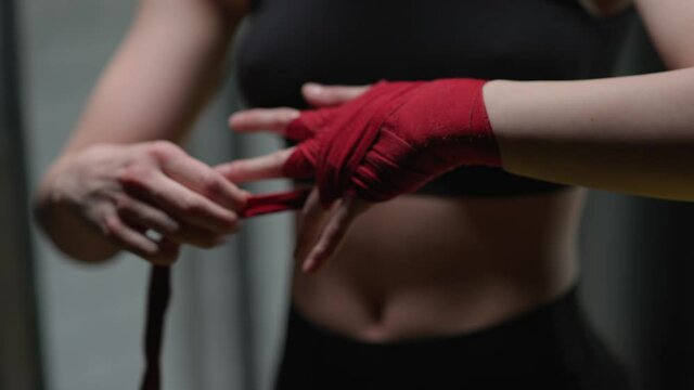 Anonymous woman fighter wraps her hands with red boxing bandages, kickboxing training day in a gym.