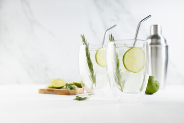 Refreshing  mojito cocktail cooking with ice cubes, lime slices, green rosemary twig blended in silver shaker, two glass mugs with straws  in elegant modern white interior on soft light wood table.