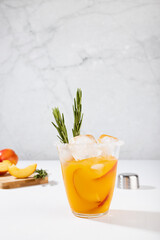 Cooking of fresh cold peach drink in glass with ice cubes, green rosemary, fruit slices, ingredients in soft light white kitchen interior, vertical.