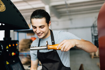 Handsome young man in apron looking at freshly roasted coffee beans while standing by coffee roasting machine.