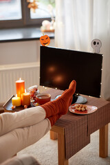 halloween, holidays and leisure concept - young woman watching tv and resting her feet on table with burning candles, cup of hot chocolate and waffle at cozy home