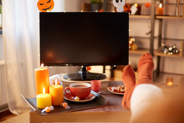halloween, holidays and leisure concept - young woman watching tv and resting her feet on table...