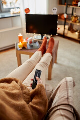 halloween, holidays and leisure concept - young woman with remote control watching tv and resting her feet on table at cozy home