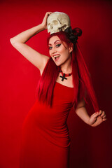 Informal Asian woman in a red evening dress with a skull in her hands. Modern witch with red hair. Halloween look