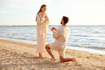 love and people concept - smiling young man with engagement ring making proposal to happy woman on beach - obrazy, fototapety, plakaty
