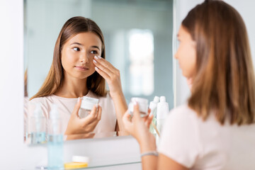 Obraz na płótnie Canvas beauty, hygiene and people concept - teenage girl applying moisturizing cream looking in mirror at bathroom