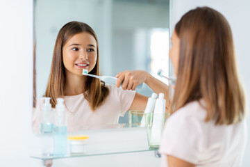 dental care, hygiene and people concept - happy smiling teenage girl with electric toothbrush brushing teeth at bathroom