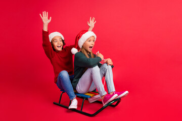 Photo of sweet excited schoolkids dressed sweaters riding sledges rising hands looking empty space isolated red color background