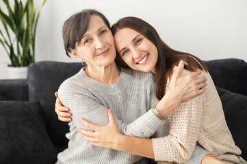 A portrait of a senior mother and an adult daughter sitting on the comfortable couch, a mom and a daughter look at camera and smile in embrace. Family ties