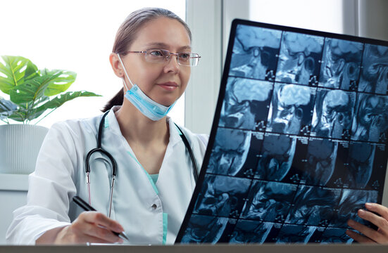 Caucasian female doctor with glasses sits at her desk and examines X-ray picture in medical clinic. Radiologist analyzes CT scan of part of human head and records result.
