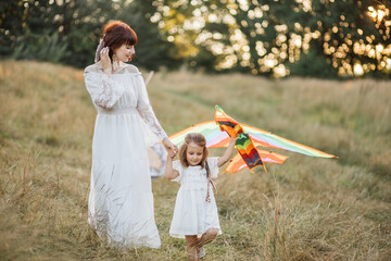 Naklejka na ściany i meble Happy family, holiday, mother's day concept. Charming mother and her cute adorable little daughter, launching a kite on nature at sunset. Boho hippie mom and child playing with kite in the field