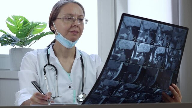 Caucasian female doctor with glasses examines X-ray picture at her desk in medical clinic. Radiologist analyses CT scan of part of human head and records result.